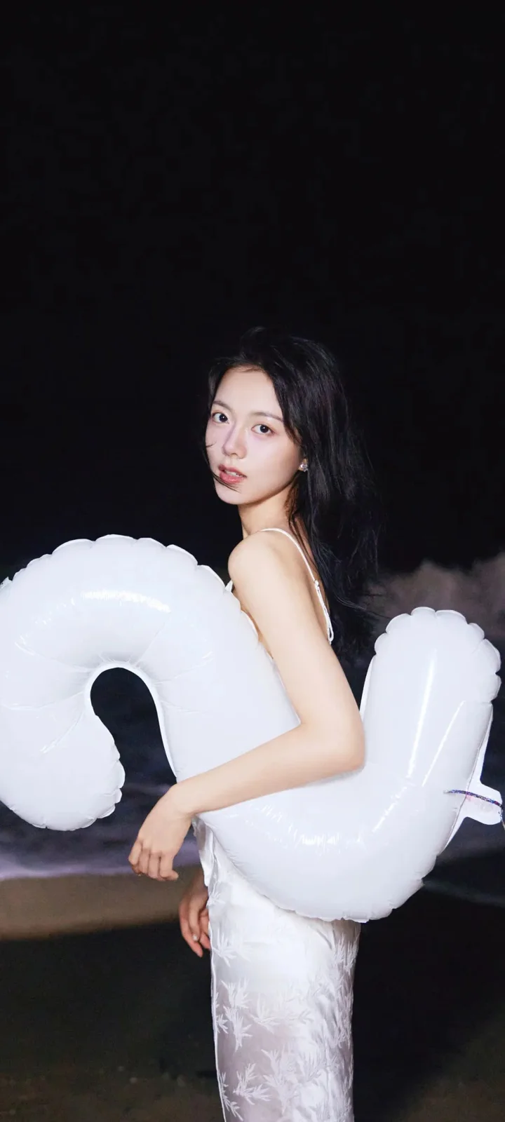 一名身穿白裙的女子在暗色背景前抱着白色充气浮具回望镜头。