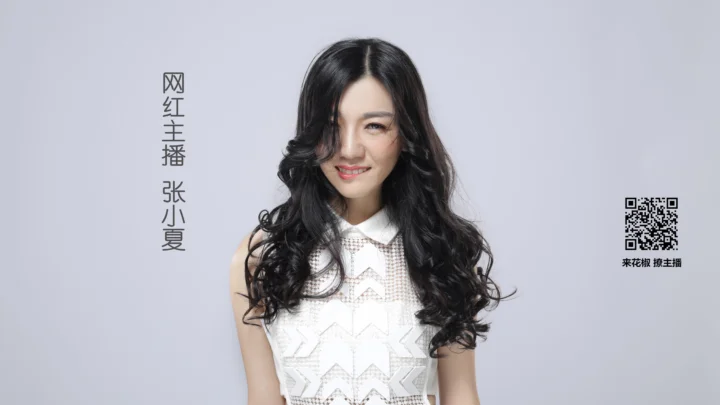 긴 검은 곱슬머리의 성인 여성이 연한 회색 배경 앞에서 흰색 민소매 상의를 입고 카메라를 향해 미소 짓고 있다.