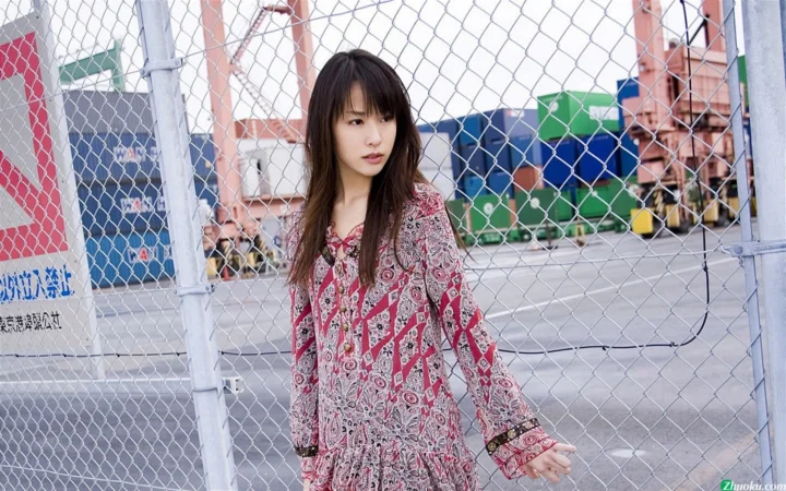 一名长发女子站在运动场围栏旁，身后是篮球场和建筑物。