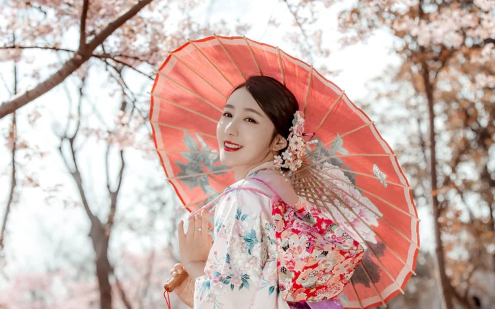 一名身穿花纹和服的女子在树下撑着红色油纸伞回眸微笑。