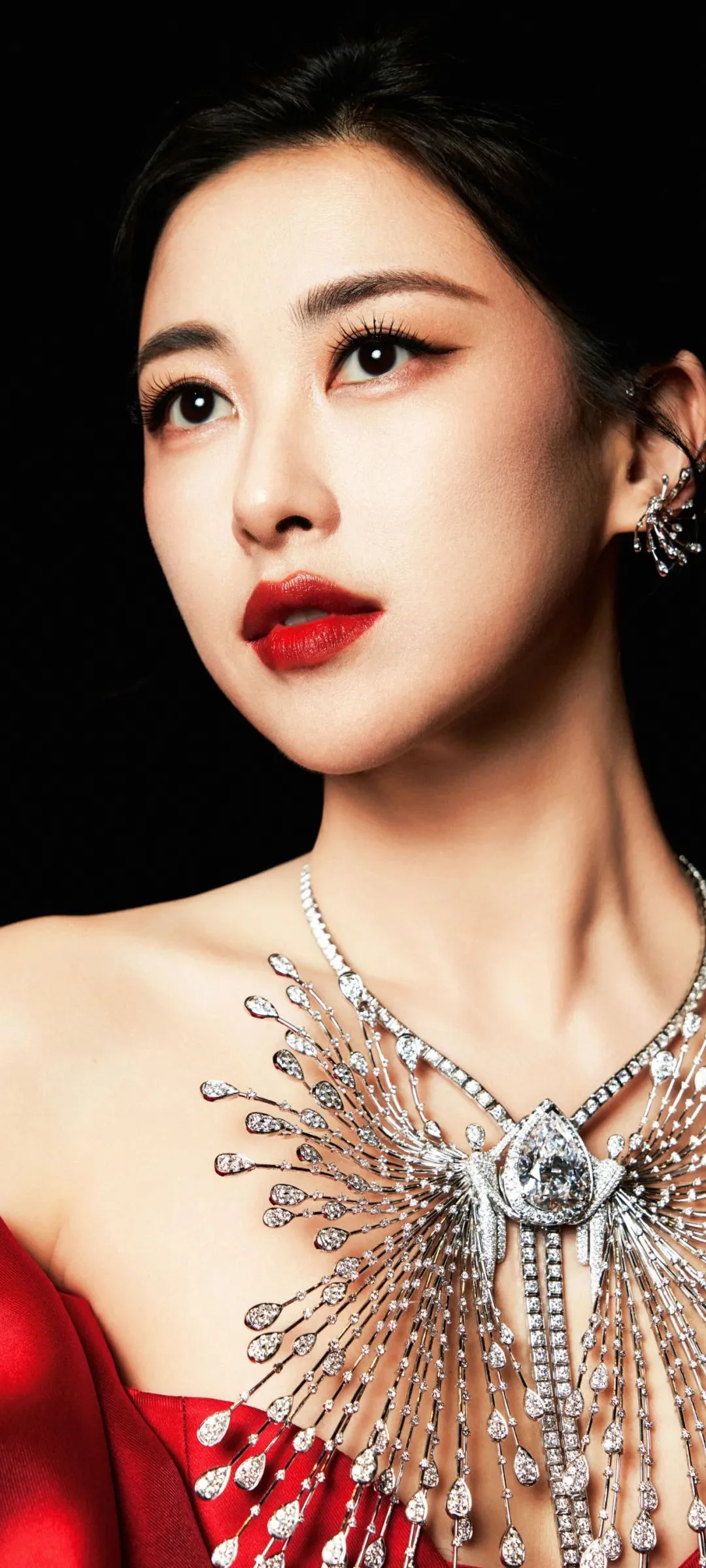 검은 배경 앞에서 빨간 드레스와 화려한 주얼리를 착용한 성인 여성을 담은 클로즈업 초상화입니다.