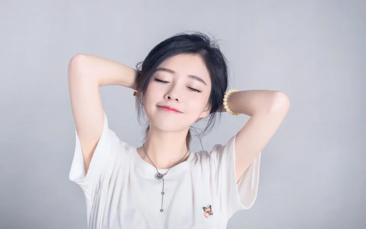 一位身穿白色上衣的女性闭着眼睛微笑，双手抬至脑后，背景为浅灰色。