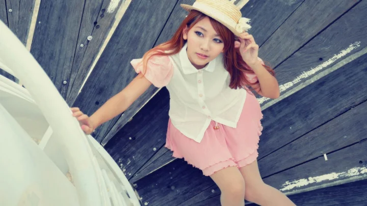 밀짚모자를 쓴 여성이 나무 데크에 서서 분홍색 옷을 입고 카메라를 바라보고 있다.