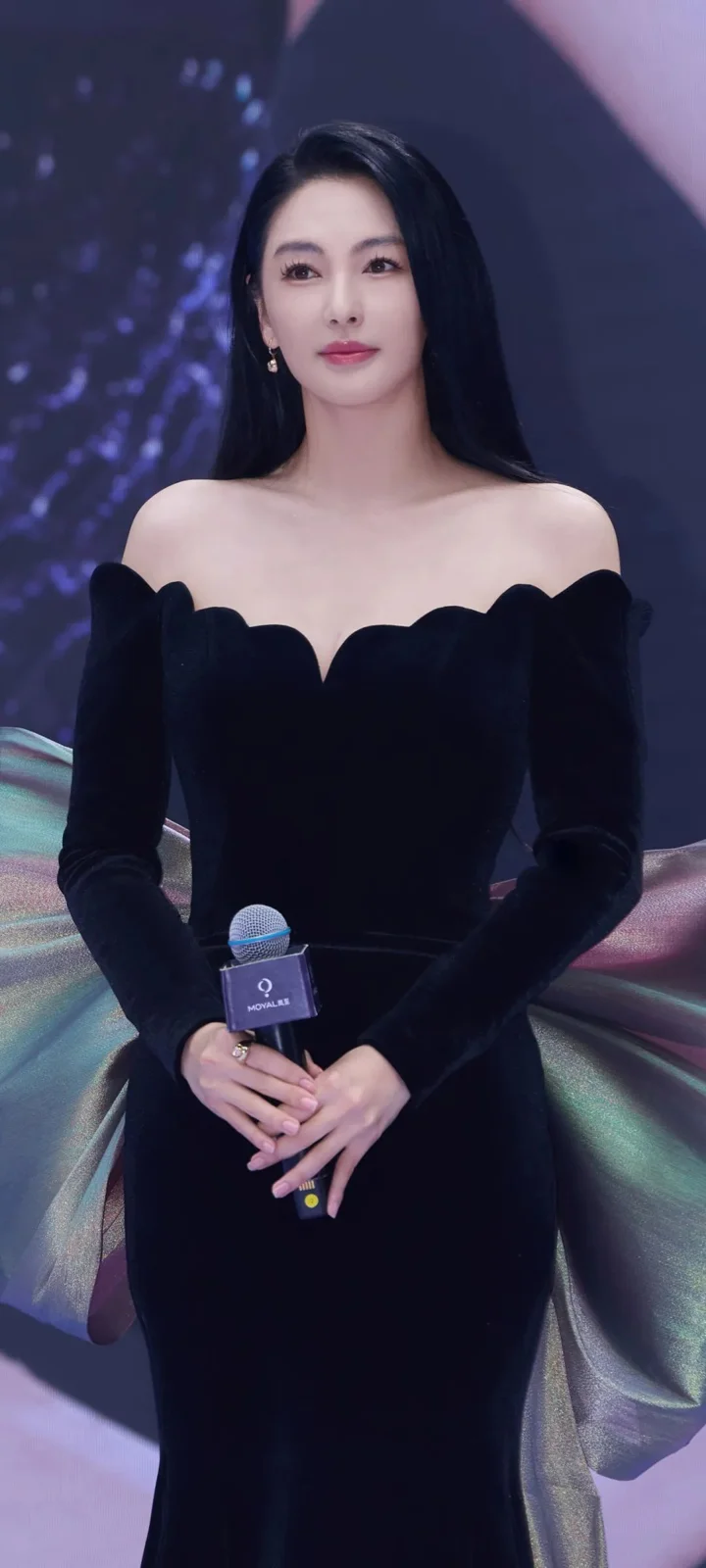 一名成年女性穿着黑色长裙，手持麦克风站在舞台背景前。
