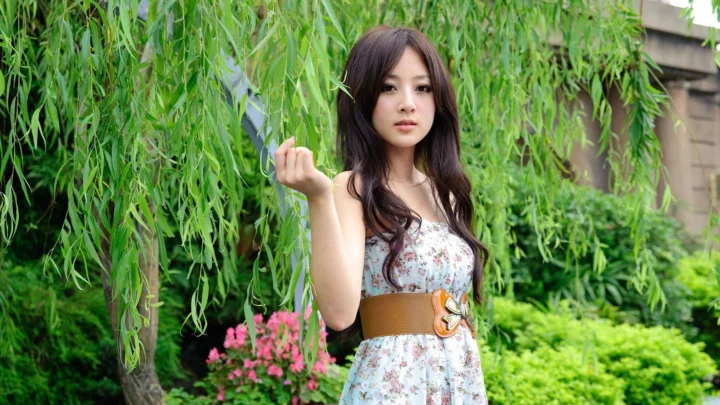 꽃무늬 원피스를 입은 성인 여성이 푸른 정원의 버드나무 곁에 서 있습니다.