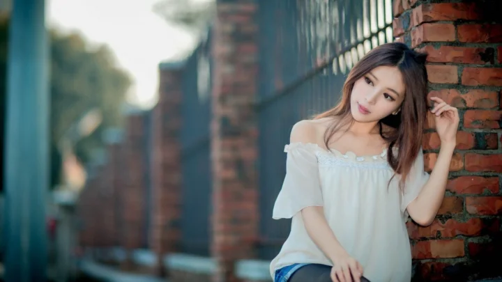 一名成年女子穿着白色露肩上衣，靠在户外砖墙旁的铁栅栏边。