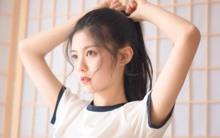 밝은 격자무늬 배경 앞에서 여성이 흰색 반팔 셔츠를 입고 머리를 묶고 있다.