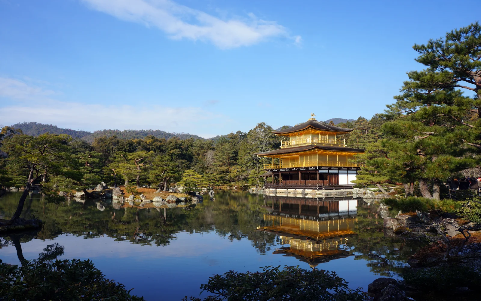 青空の下、湖畔の金色の楼閣と木々が水面に映る穏やかな風景です。