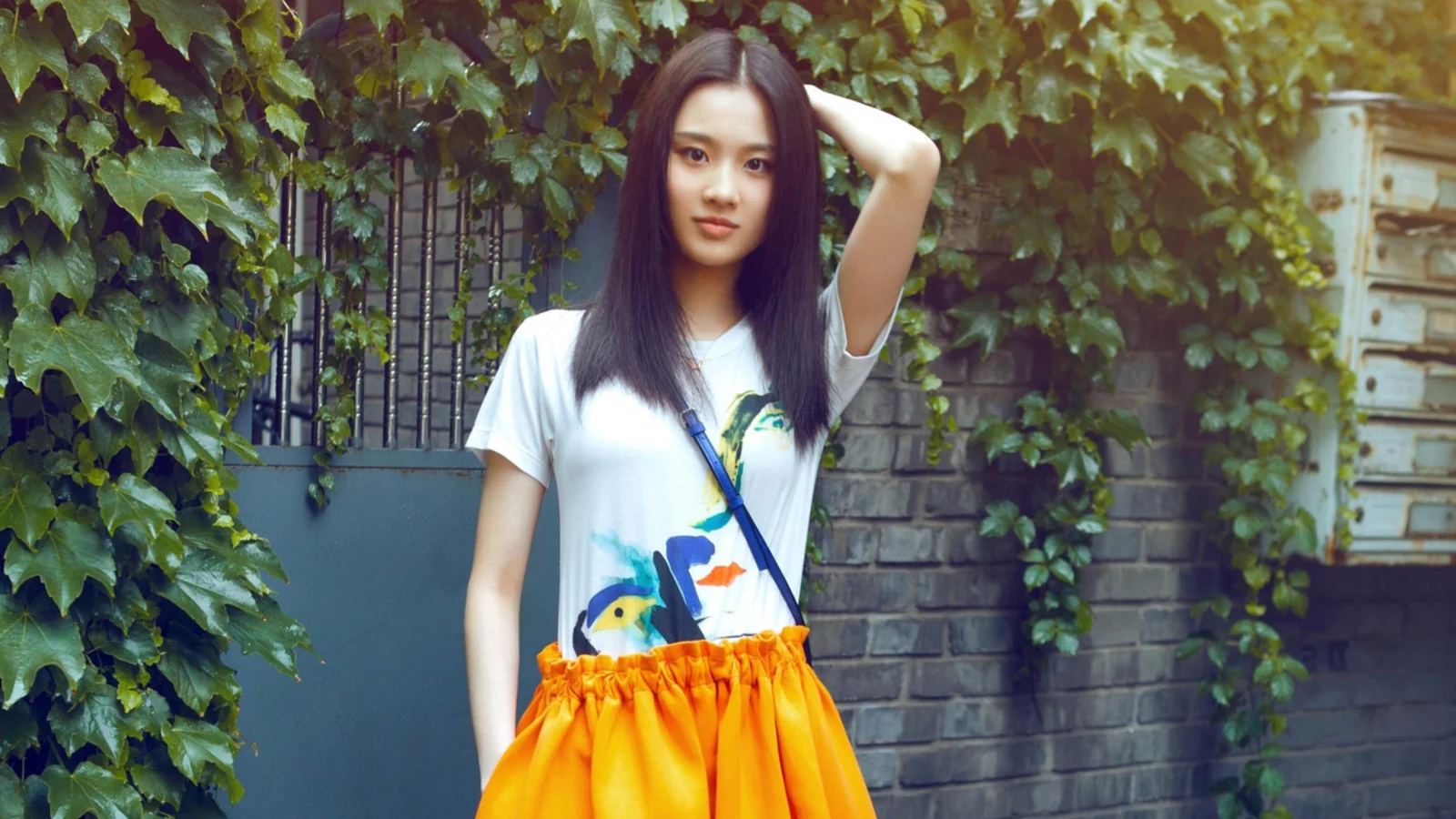 긴 머리의 여성이 담쟁이덩굴로 덮인 벽돌墙 앞에 서서 흰색 상의와 주황색 치마를 입고 있습니다.