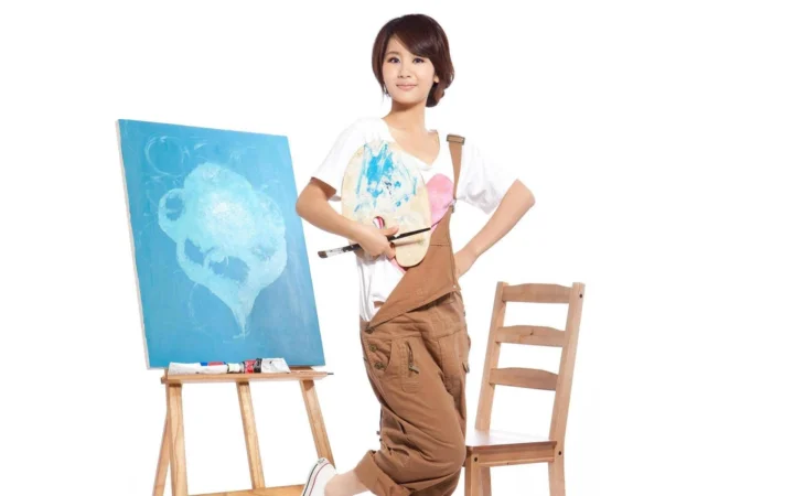 一名成年女性站在画架和木椅旁，手持画笔，白色背景前陈列着一幅蓝色画作。