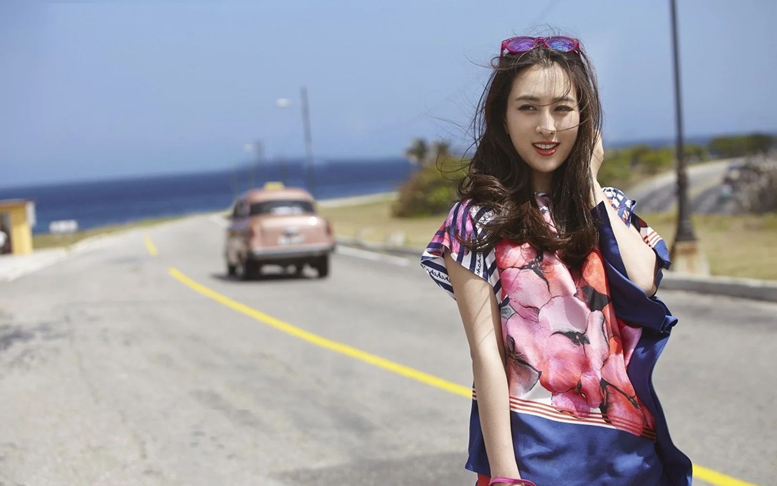 一名女子站在海邊公路旁，遠處可見海洋和一輛行駛中的汽車。