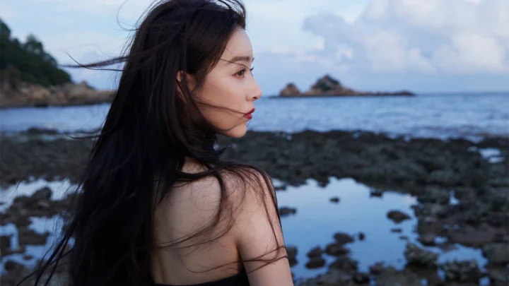 一名成年女性站在海边岩石岸旁，侧脸望向平静的海面。