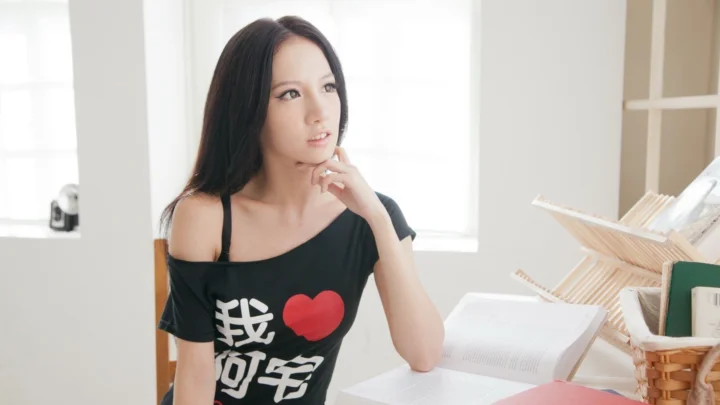 一名成年女性坐在明亮的窗边，手托下巴，身旁放着打开的书本和收纳篮。