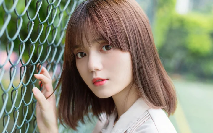 一名棕色长发女性站在铁丝网旁，望向镜头，背景是虚化的绿色户外景色。