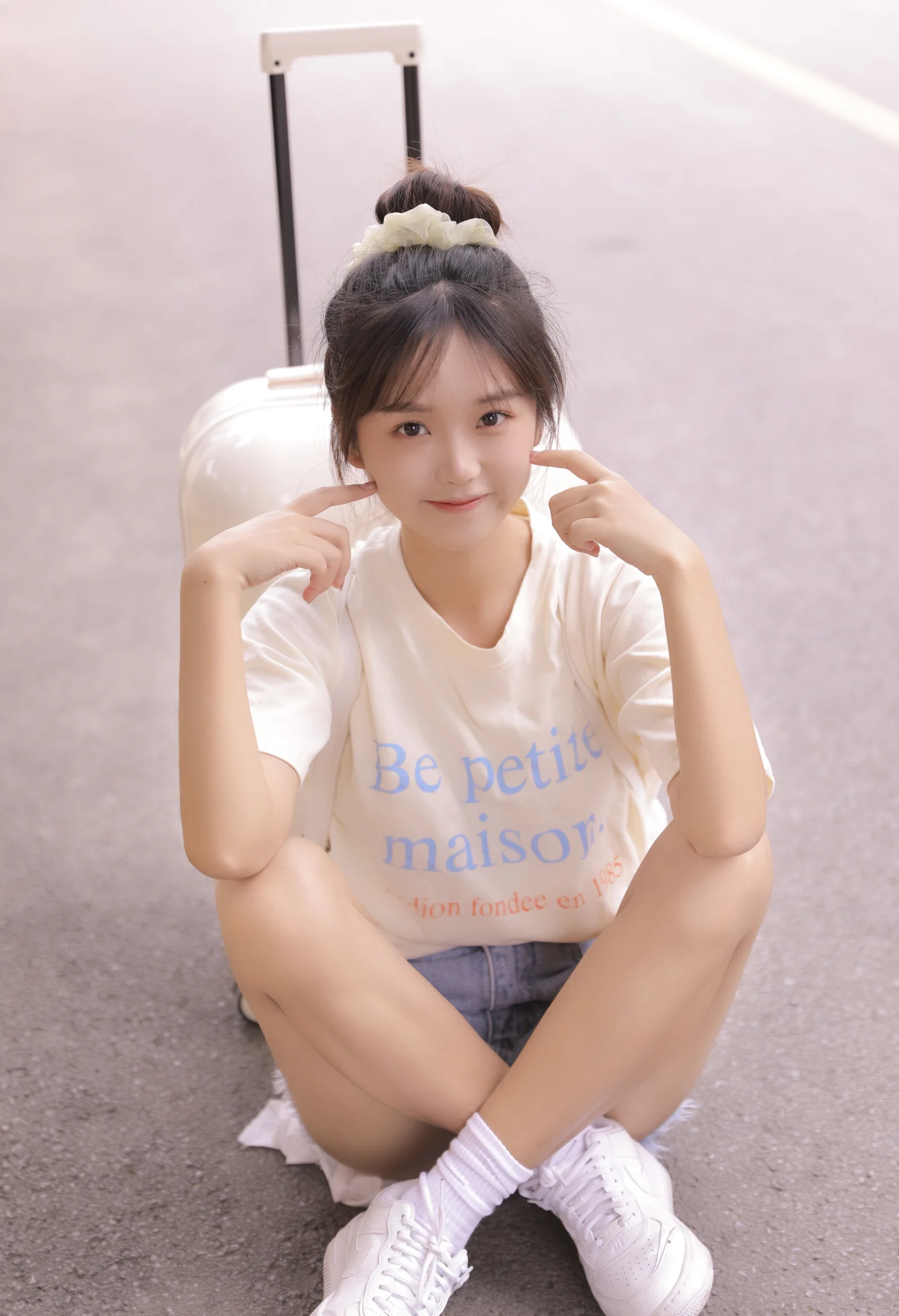 一名扎着发髻的年轻女子坐在地面上，身后放着一个白色旅行箱。
