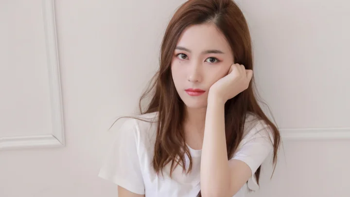 갈색 긴 머리의 여성이 흰색 반팔 셔츠를 입고 밝은 색 벽 앞에서 손으로 뺨을 받치고 있다.