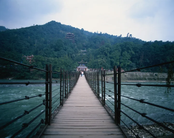 一座木制悬索桥横跨湍急河流，通向青山密林中的建筑群。