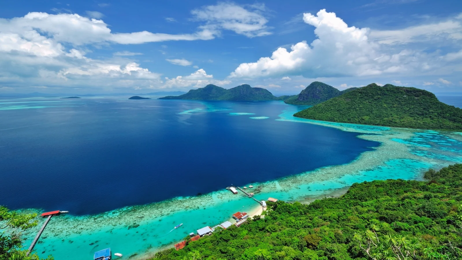 航拍视角下，湛蓝海水、绿色群岛、珊瑚礁与晴朗天空构成壮丽的海洋风景。