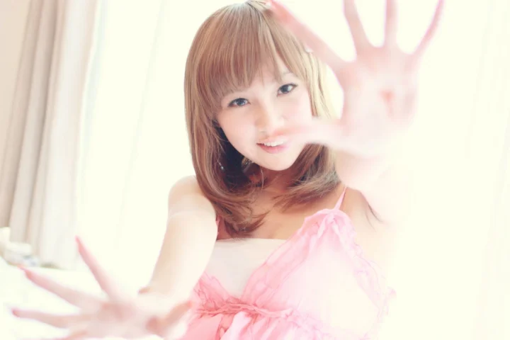 갈색 머리의 여성이 분홍색 원피스를 입고 밝은 빛 속에서 양손을 카메라 쪽으로 뻗고 있다.