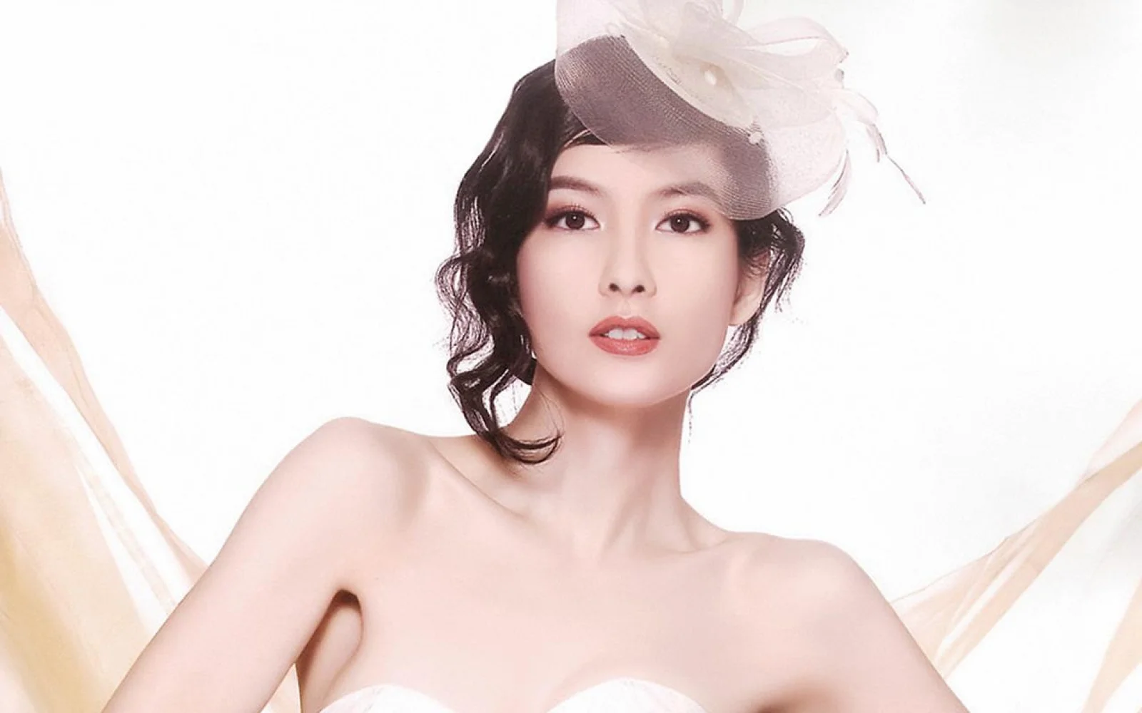 一名戴着白色装饰帽、穿着无肩带白色服装的女性站在明亮的白色背景前。