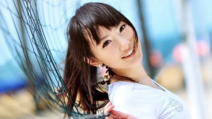짙은색 긴 머리의 여성이 흰색 상의를 입고 그물 구조물 옆에서 밝게 미소 짓고 있습니다.