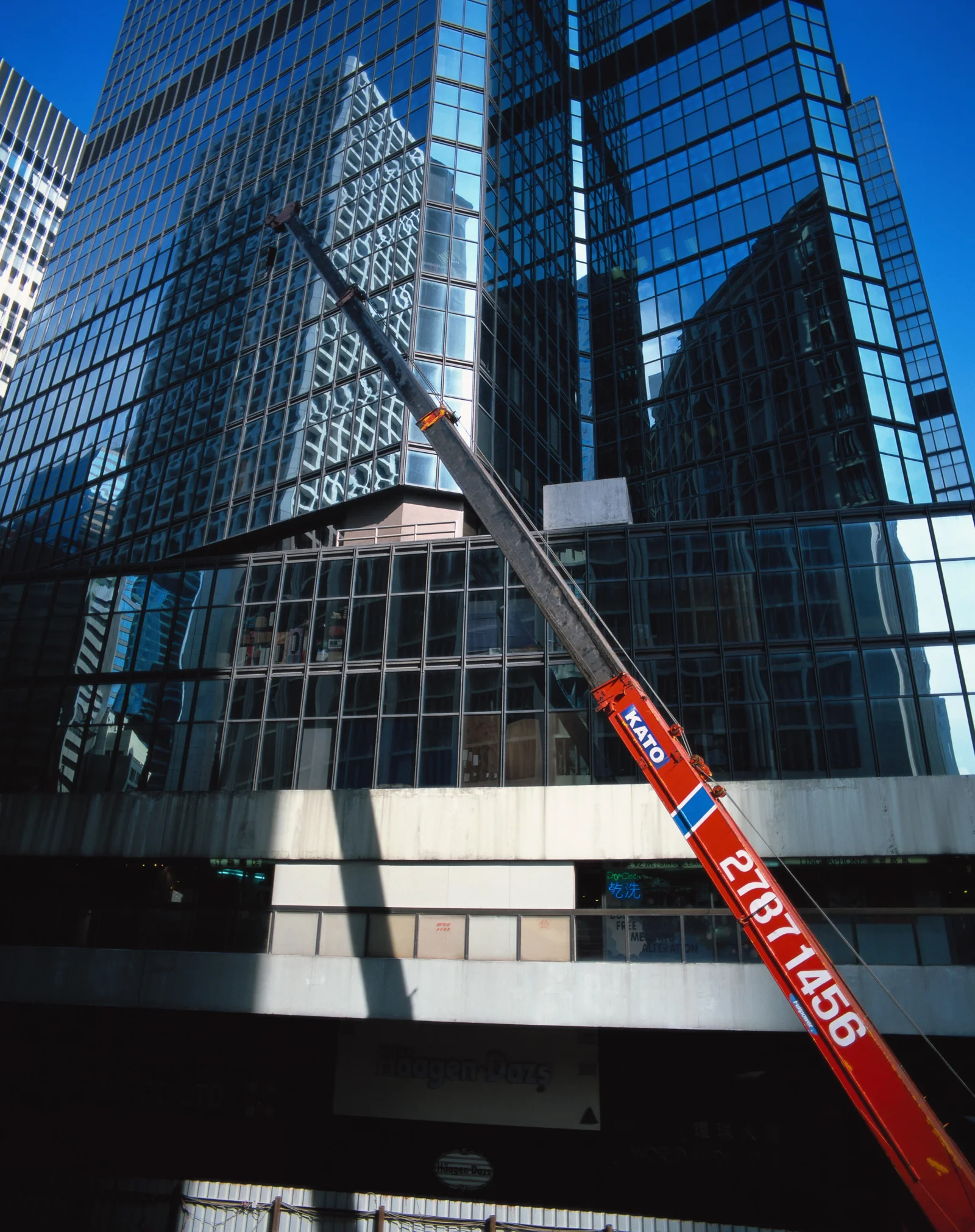 高耸的玻璃幕墙建筑在蓝天下映照出城市景观，一台红色施工吊臂斜穿画面。