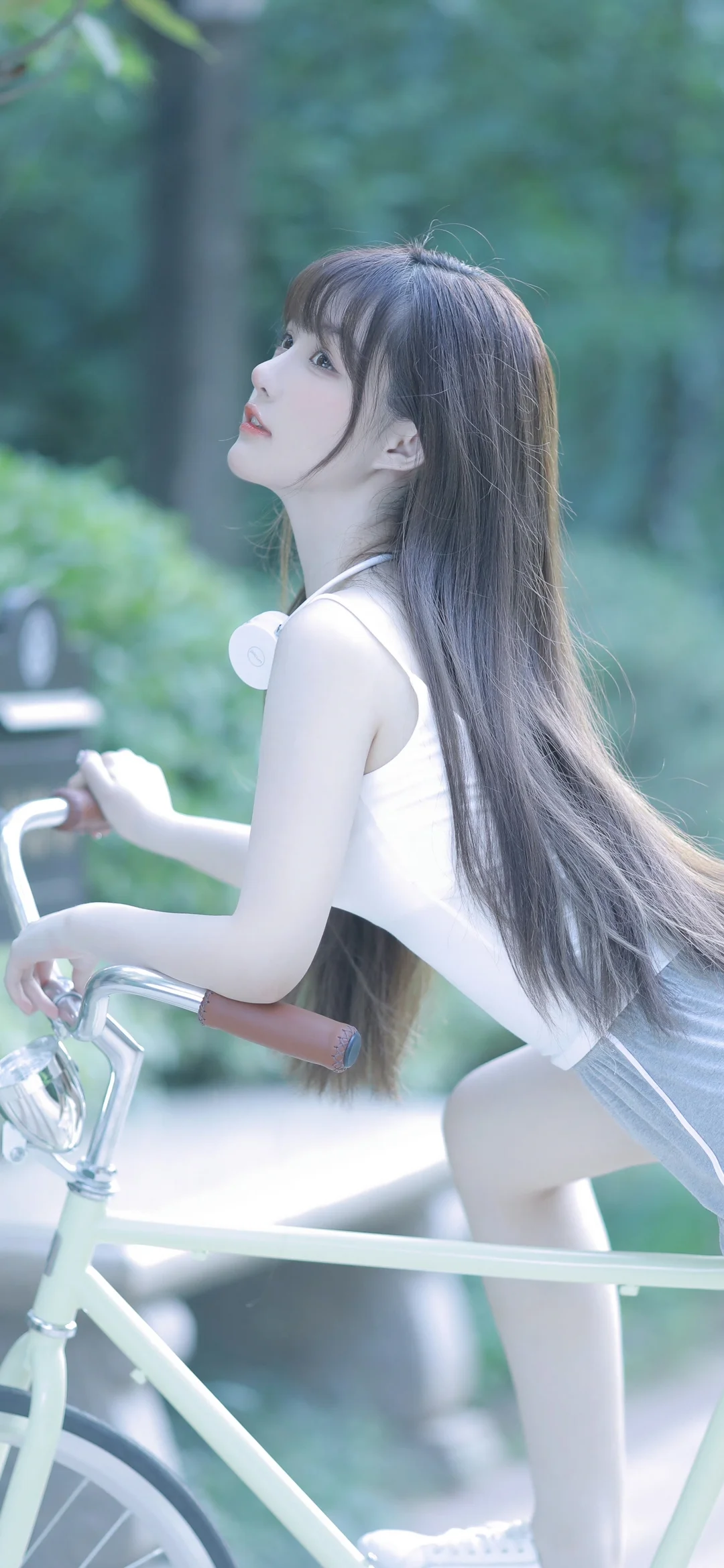 一名長髮女性坐在白色自行車上，身後是虛化的綠色樹林。