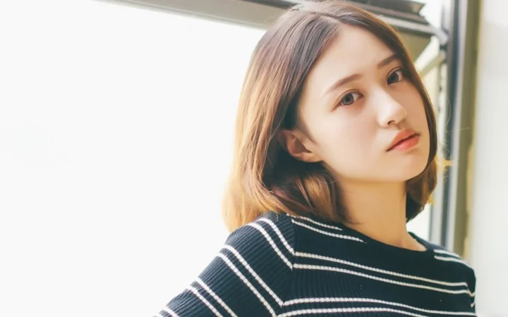 一名棕色短发女性穿着黑色条纹上衣站在窗边，面向镜头。