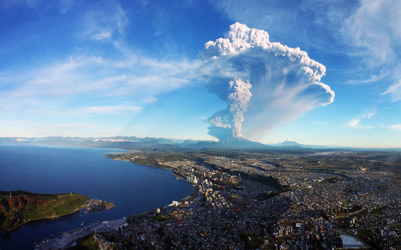 航拍画面展示了海湾城市、远处火山和高耸云柱。