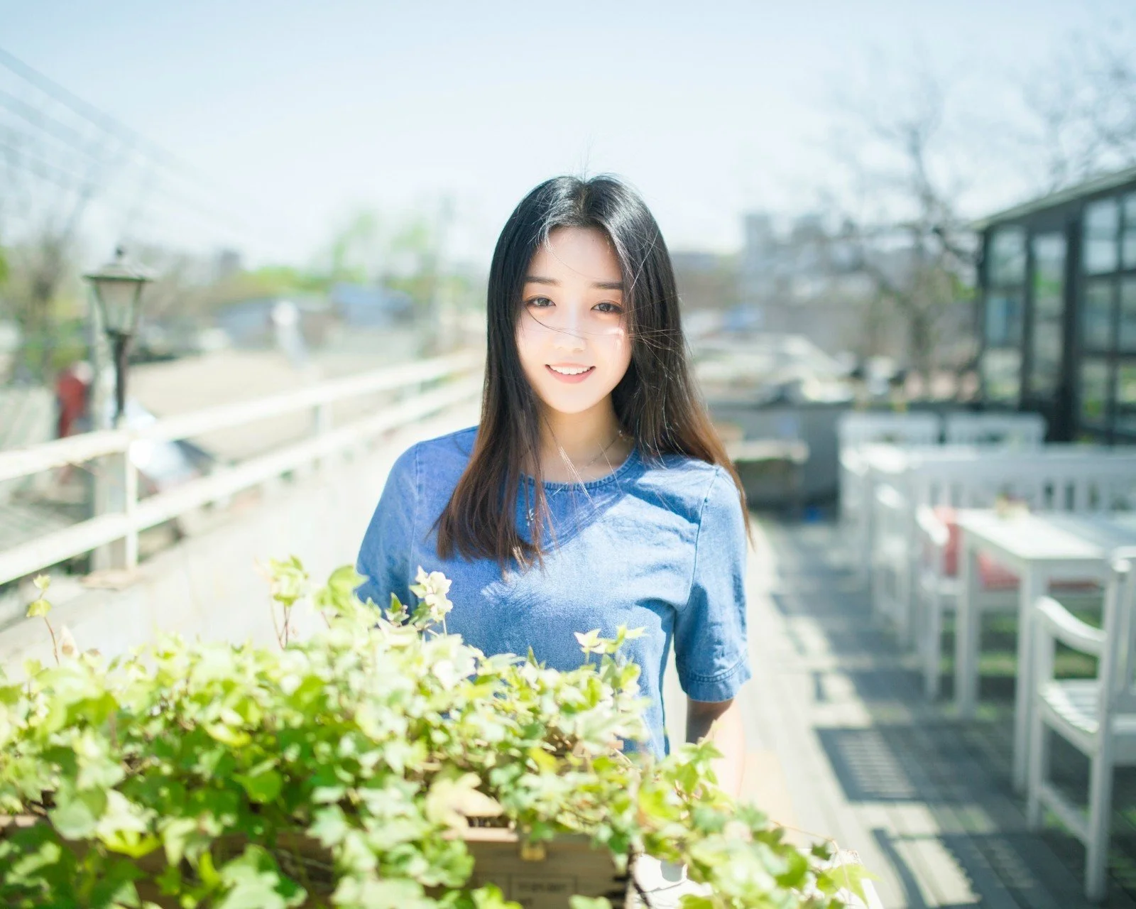青いトップスを着た女性が、手前の鉢植えに囲まれた明るいバルコニーに立っています。