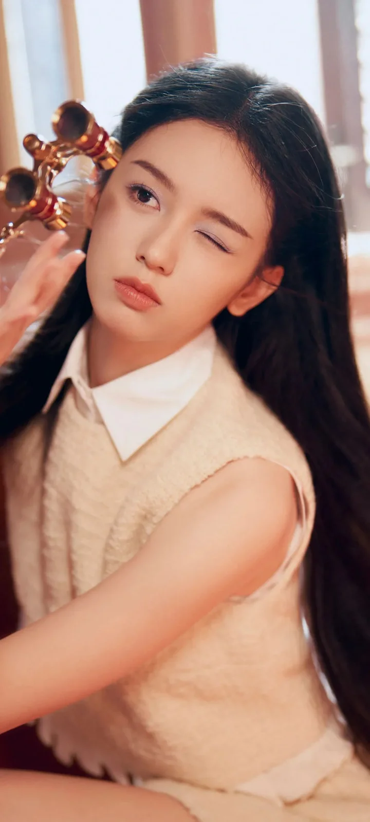 一名长发女子穿着米色无袖针织衫，对镜头眨眼。
