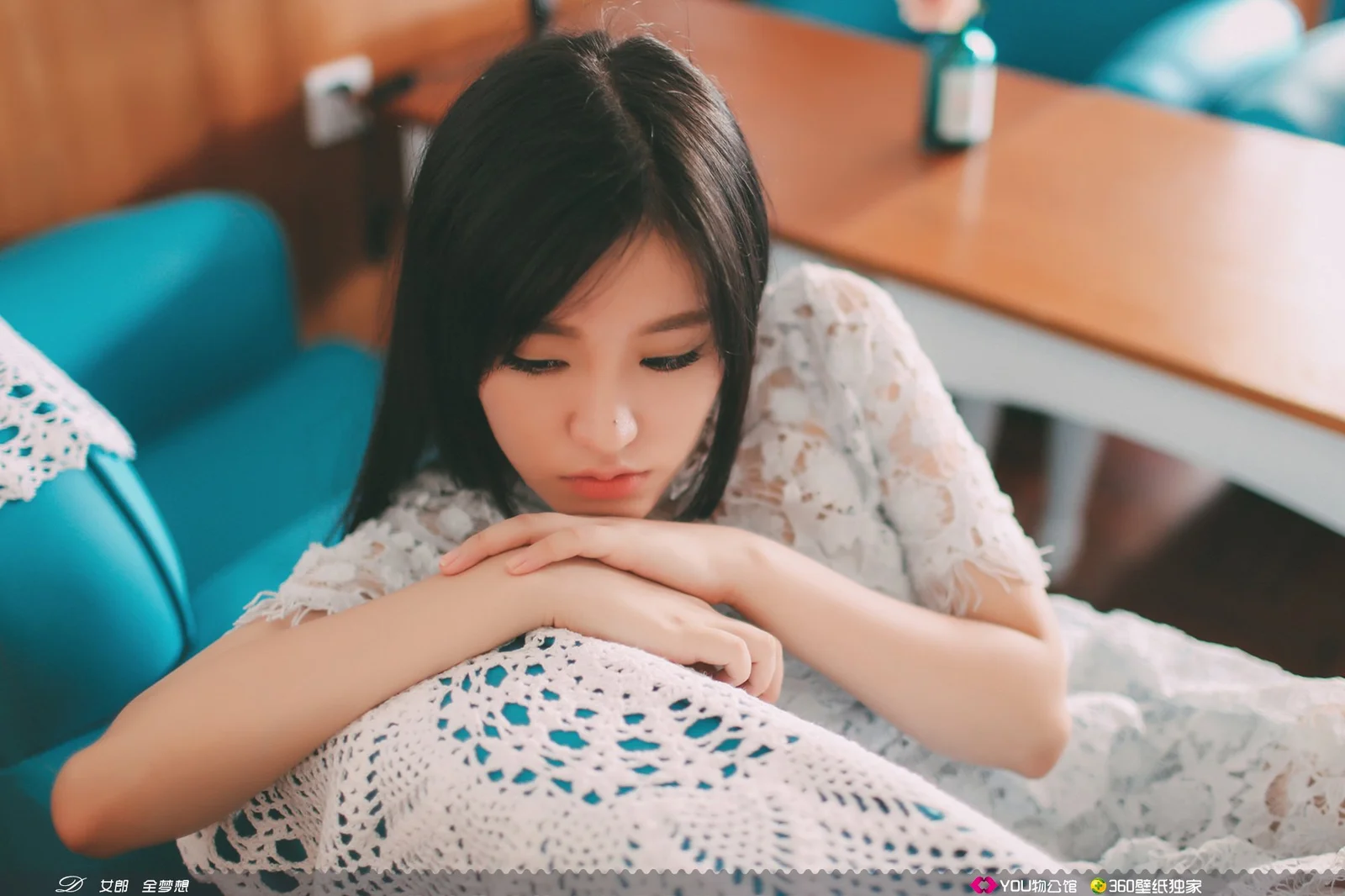 一名黑发女性身穿白色蕾丝服装，俯身靠在桌边的白色钩针织物上。