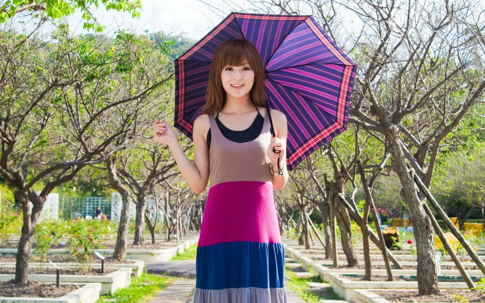 一名成年女子在树木环绕的公园小径上撑着条纹雨伞。