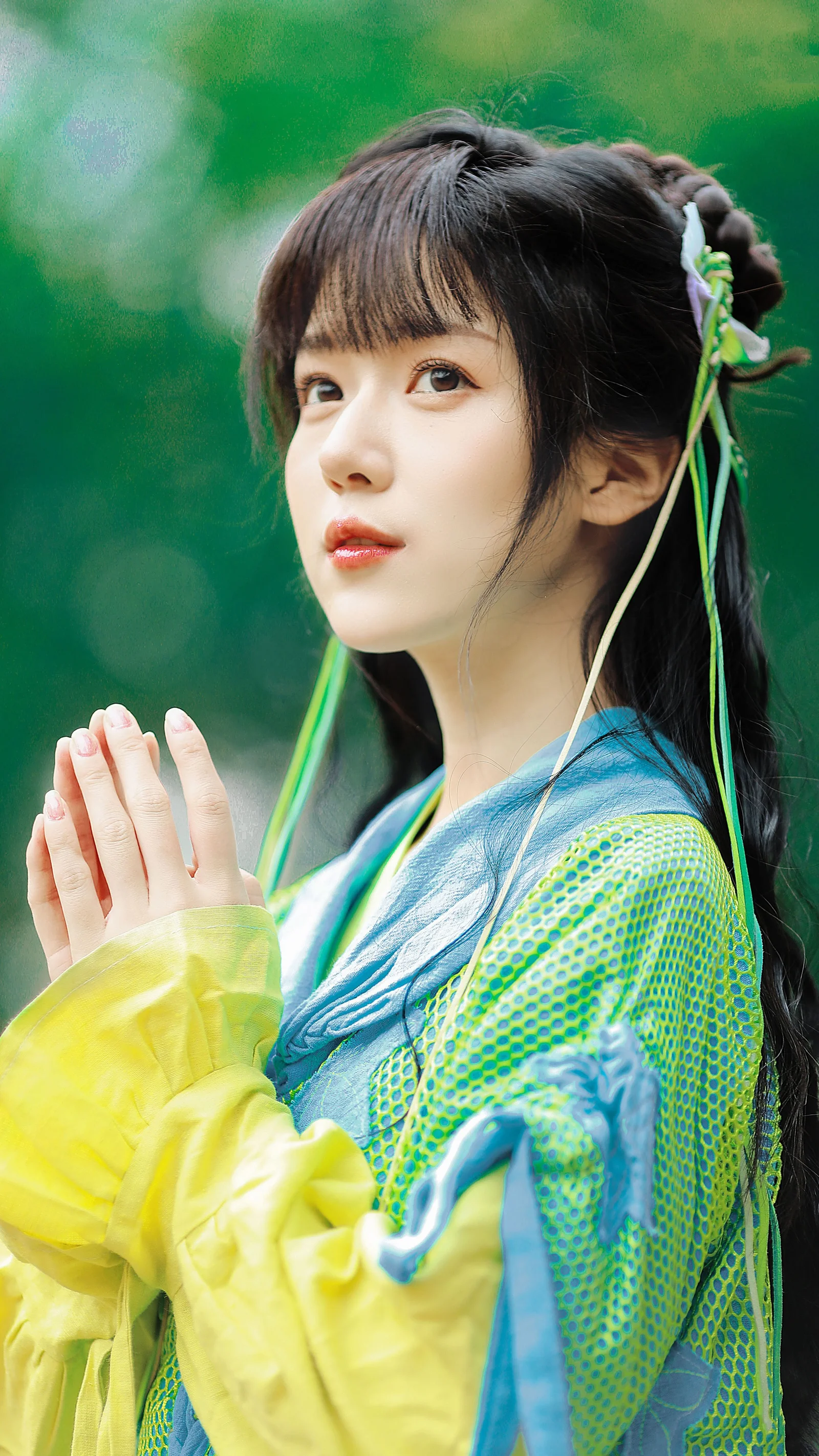 검은 머리의 여성이 선명한 청록색과 노란색 옷을 입고 초록색 배경 앞에서 두 손을 모은 채 카메라를 바라보고 있습니다.