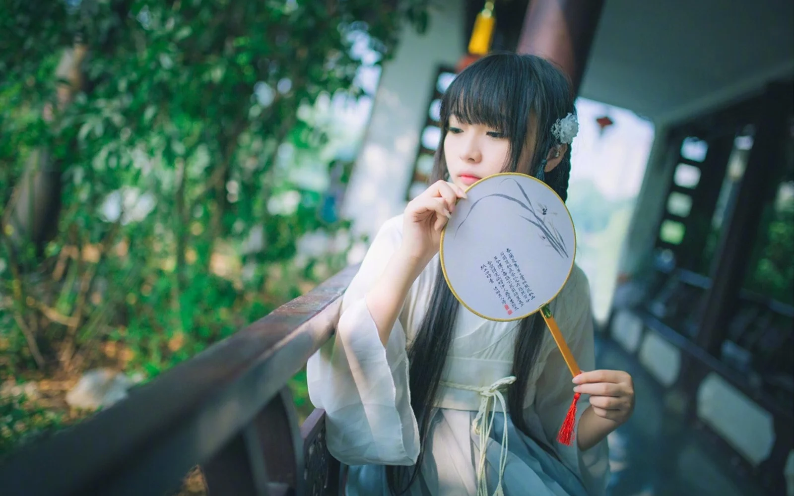 一名身穿浅色传统服饰的女子站在木栏旁，手持团扇望向镜头。