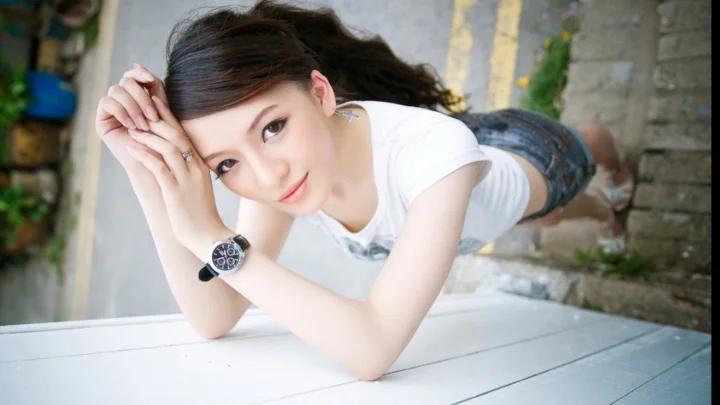 一名成年女性身穿白色上衣和牛仔短裤，俯身靠在户外桌面上看向镜头。