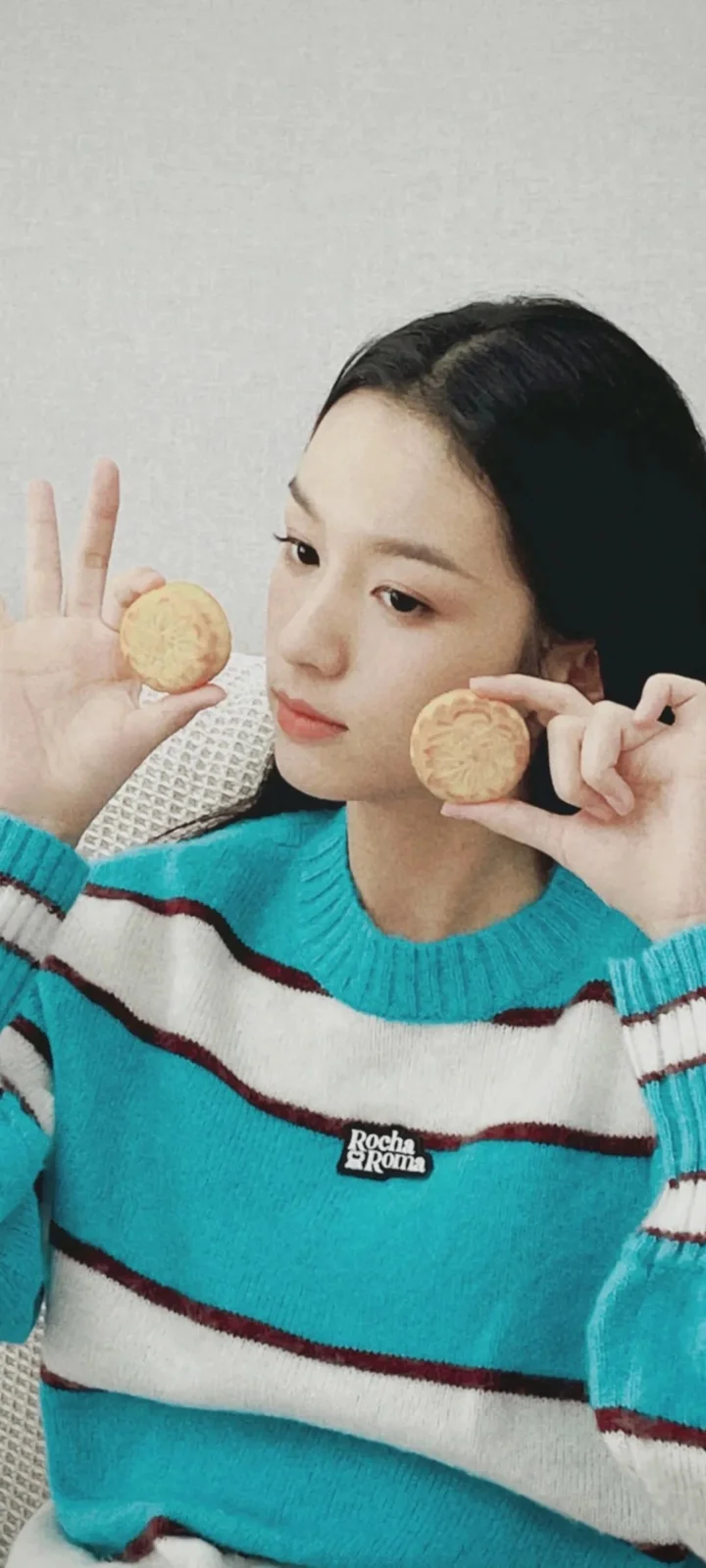 一名女子穿着蓝绿色条纹毛衣，手持两块圆形饼干靠近脸颊。