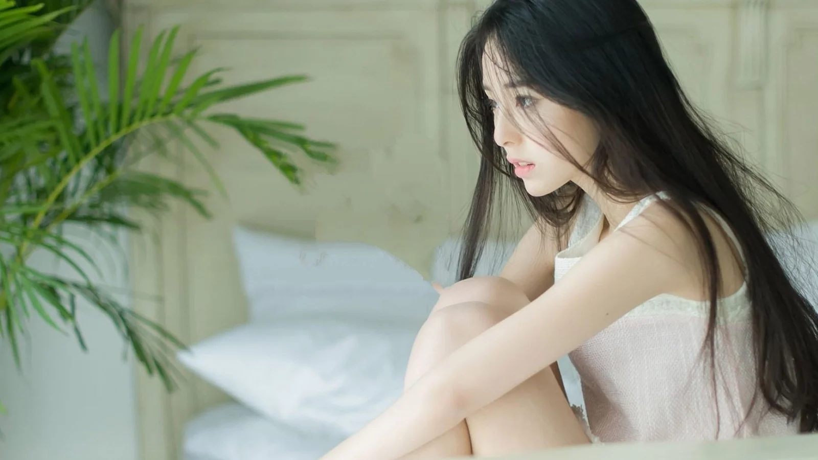 一名长发女子坐在明亮的室内床边，旁边有一株绿植。