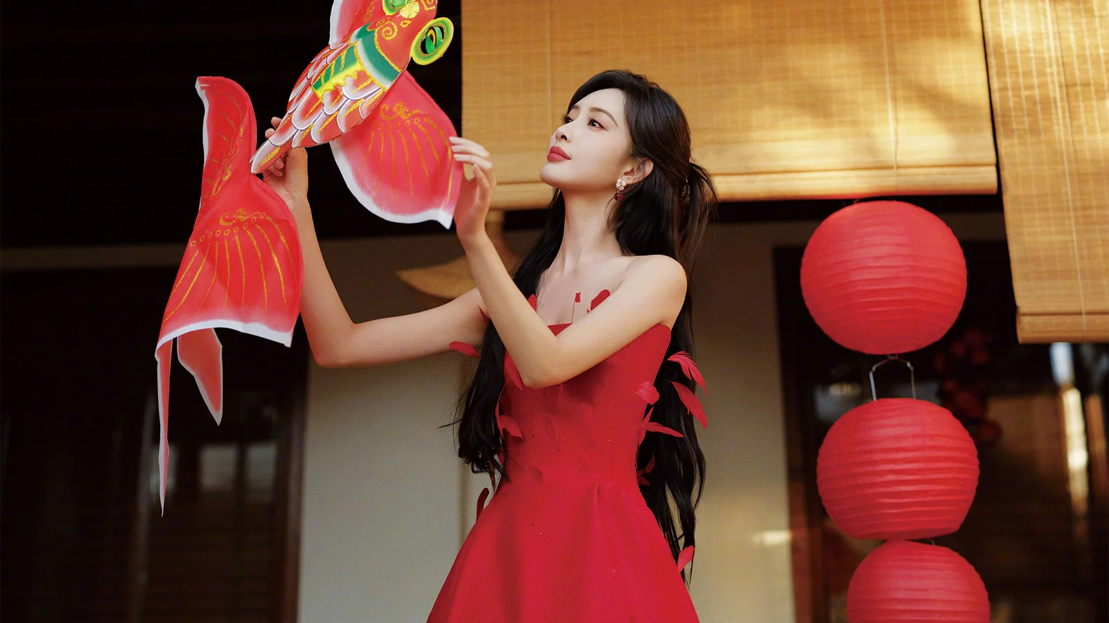 一名穿红色连衣裙的成年女子站在室内红色灯笼旁，手持彩色装饰物。