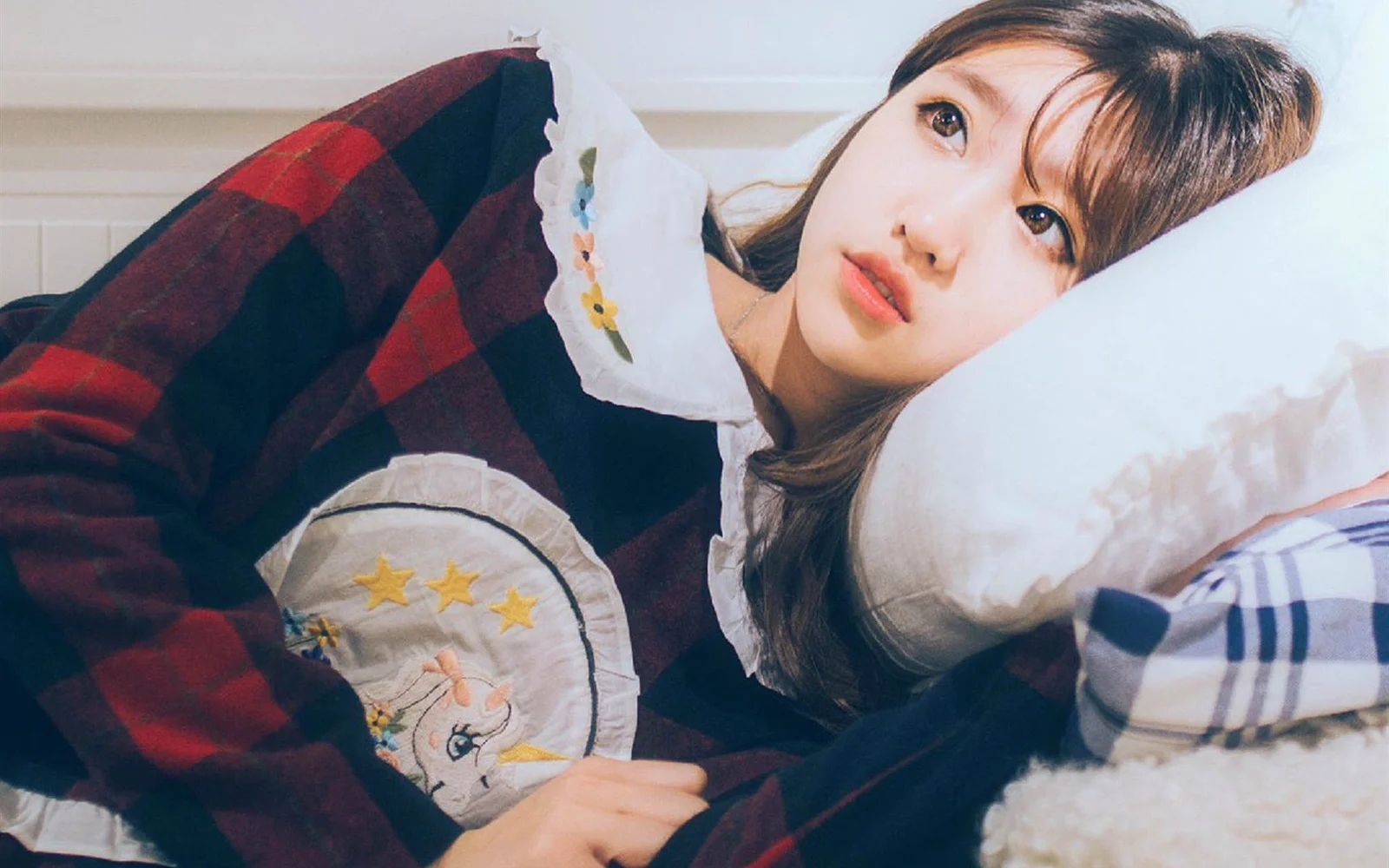 一名女性穿着格纹上衣侧躺在床上，头靠着白色枕头看向镜头。