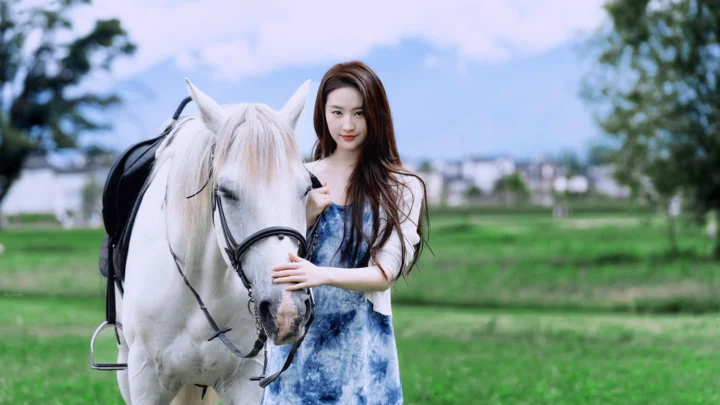 一名女子站在绿地上，身旁是一匹白马，远处可见树木和低矮建筑。