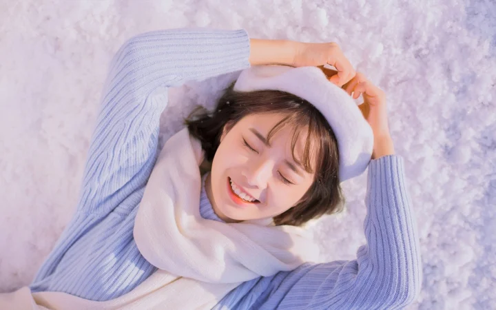 白い冬帽子とマフラーを身につけた女性が、淡いラベンダー色の雪景色のような背景で目を閉じて微笑んでいる。