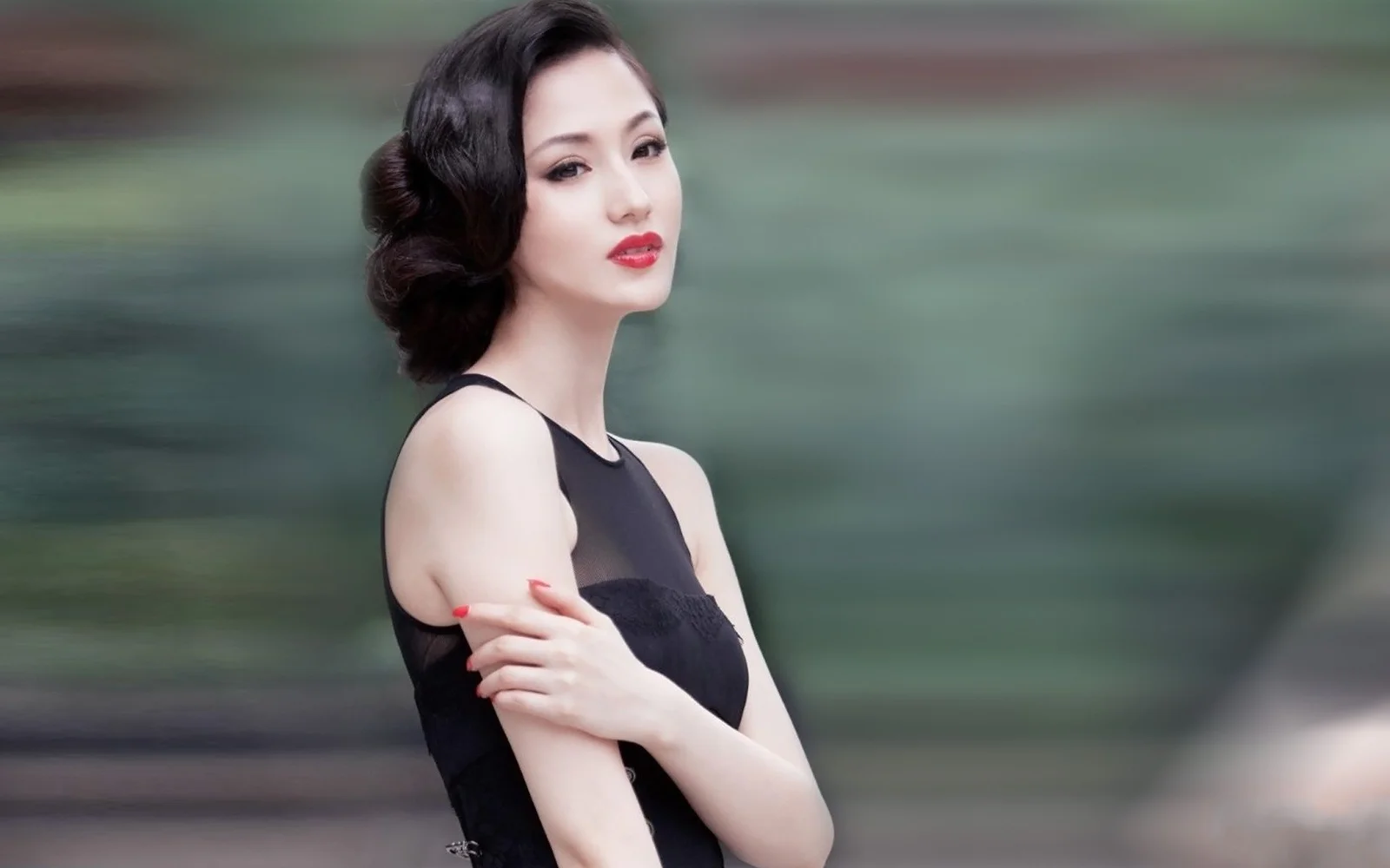一位身穿黑色连衣裙的成年女性在模糊的绿色背景前交叉双臂。