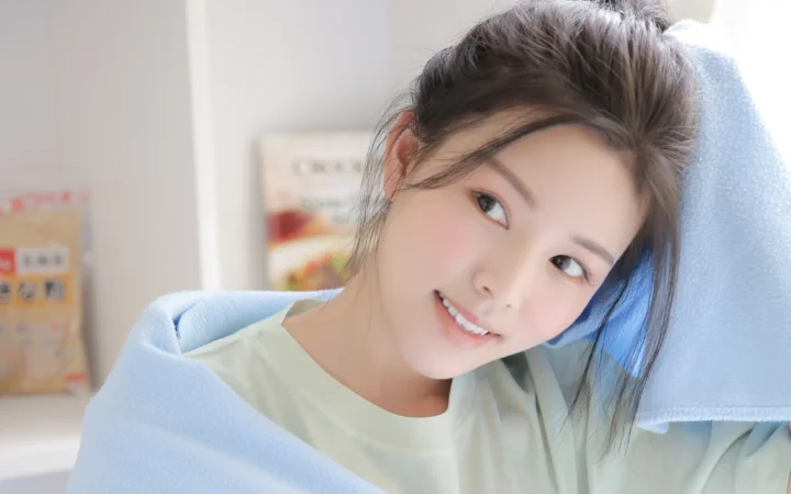 긴 머리의 여성이 실내에서 연한 파란색 수건에 기대어 카메라를 향해 미소 짓고 있다.