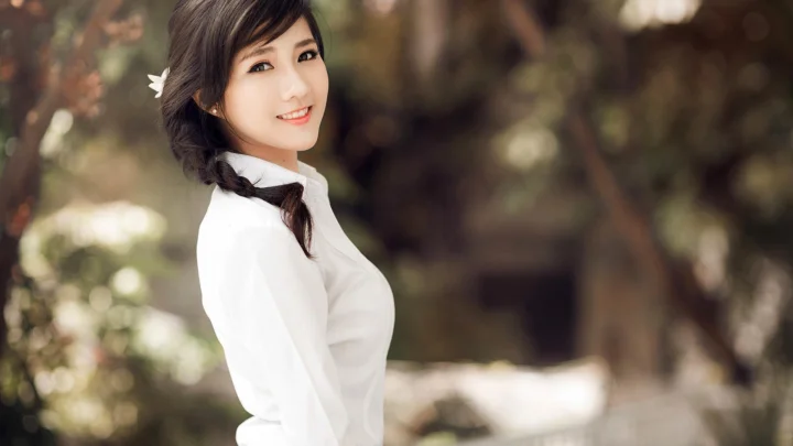 一位身穿白色上衣、留着编发的女性站在虚化的户外树林中。