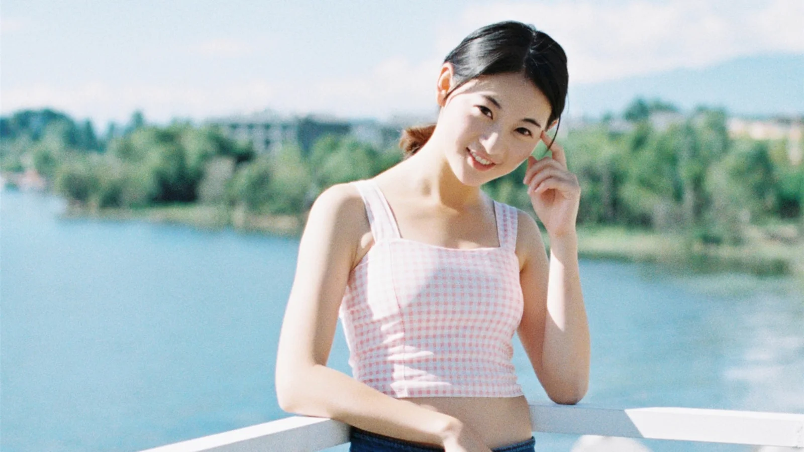 一名年轻女子站在湖畔栏杆旁微笑，身后是宁静的湖水与绿树。