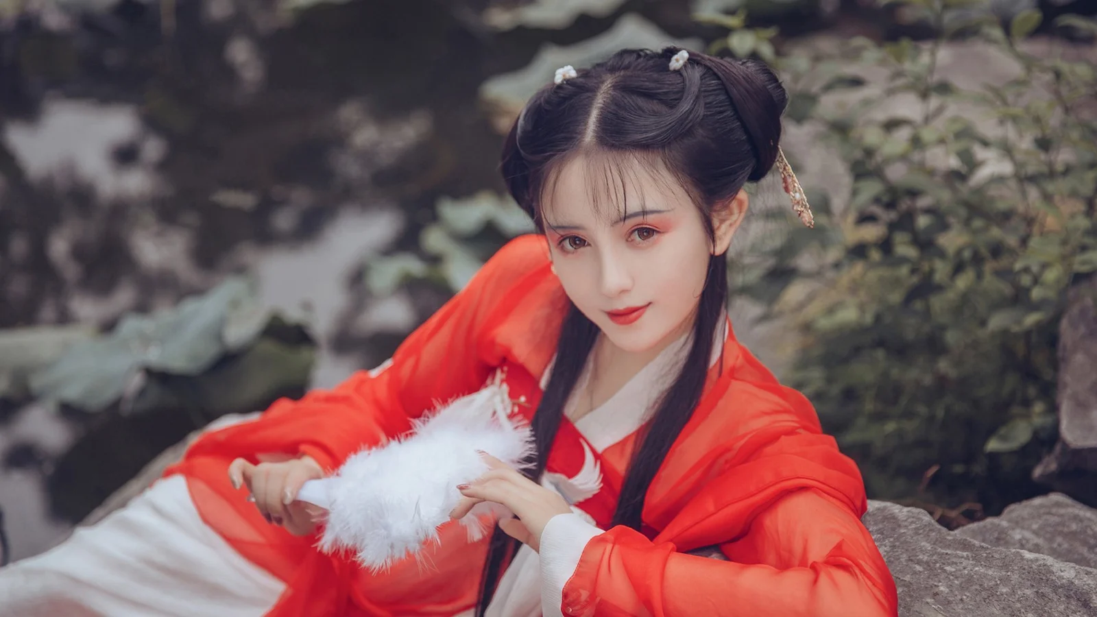 一名身穿红色传统风格服饰的女性坐在户外岩石旁，手持白色羽毛扇。
