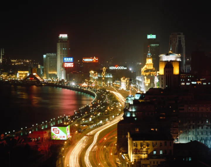 夜色中的城市滨水天际线灯火璀璨，沿岸道路延伸出明亮的车流光轨。