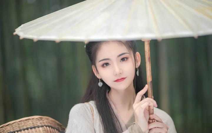 一名女子手持浅色伞站在深绿色背景前。