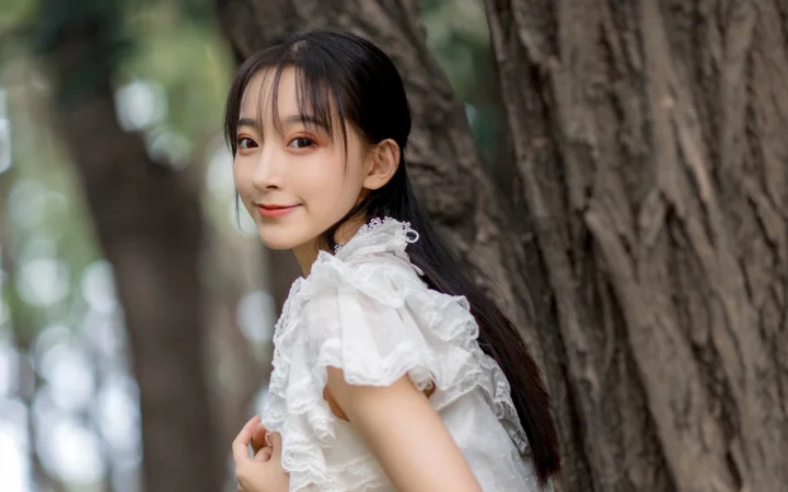 一名黑色长发女子身穿白色褶边上衣站在树旁，回头面向镜头微笑。